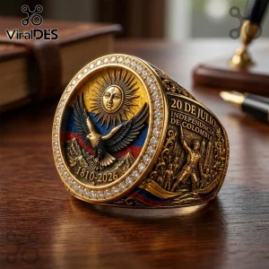 Gold Colombian independence ring with eagle, sun, and 1810-2026 text surrounded by clear embedded stones on wood desk.!