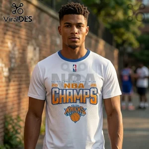 Man wearing white New York Knicks 2026 NBA Champs graphic t-shirt with blue collar trim against a brick wall background.!