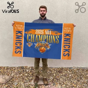 Man holding a blue and orange New York Knicks 2026 NBA Champions wall flag with team logo on a grey textured background.!