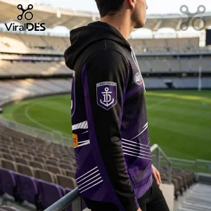 Man wearing a purple and black AFL Fremantle Dockers hoodie with an anchor crest and Est. 1984 text at a stadium.!