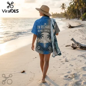 Woman wearing blue shirt with palm tree graphic and text I'M RECHARGING DO NOT CONTACT me, walking on a sandy beach.!