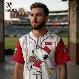 Man wearing white Peanuts baseball jersey with red pinstripes, red sleeves, and Snoopy graphics at a stadium.!