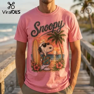 Pink t-shirt with Snoopy graphic wearing sunglasses and floral shirt next to a surfboard on a beach at sunset.!