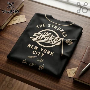 The Strokes Reality Awaits 2026 Tour black baseball jersey featuring gold band logos, text, and signatures on a wood table.