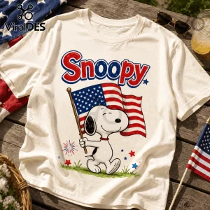White crewneck t-shirt featuring Snoopy holding an American flag with red Snoopy text and patriotic stars on a wood table.!