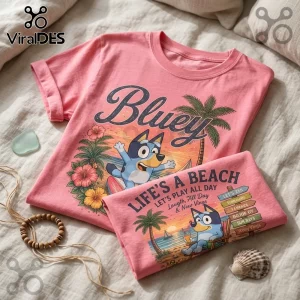 Pink crew neck t-shirt featuring Bluey cartoon character, palm trees, and Life's A Beach text on a beige linen bedspread.!