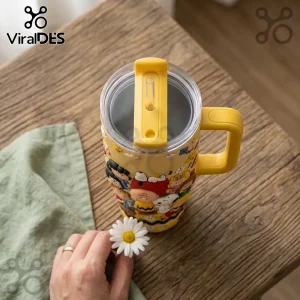 Yellow stainless steel tumbler featuring Peanuts character graphics, a yellow handle, and a clear sliding lid on wood desk.!