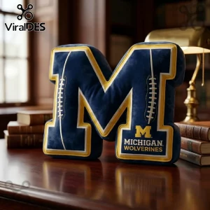 Michigan Wolverines letter M plush pillow in navy blue and yellow with embroidered white football lace details on a desk.!