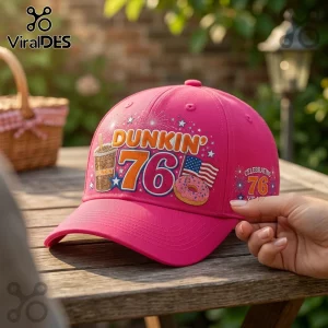 Dunkin Donuts 76th Anniversary pink baseball cap with iced coffee, donut, and flag graphics on a wooden table outdoors.
