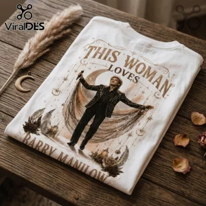 White t-shirt featuring a Barry Manilow illustration, gold crescent moon, stars, and feathers on a wooden table surface.!