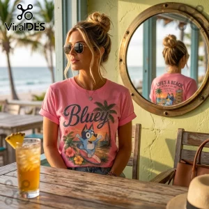 Woman wearing a pink Bluey t-shirt with tropical beach graphics, sitting at a wooden table near a round mirror.!