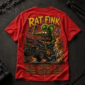 Red graphic t-shirt featuring Rat Fink driving a hot rod with fire, skulls, and automotive text on a dark workshop table.!