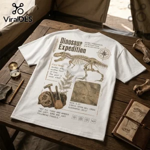 White graphic t-shirt featuring Tyrannosaurus Rex skeleton, compass, topographical map, and paleontology text on wood table.!