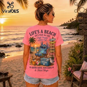 Pink t-shirt featuring a cartoon Stitch graphic, beach scene, sunset, and text Life's a Beach on a woman at the shoreline.!
