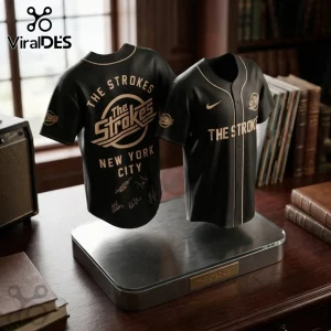 The Strokes Reality Awaits 2026 Tour black baseball jersey featuring gold graphics, NYC text, and band signatures on stand.