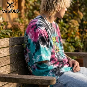 Man wearing a pink, turquoise, and black tie-dye Blink-182 band t-shirt with graffiti art, sitting on a wooden bench.!