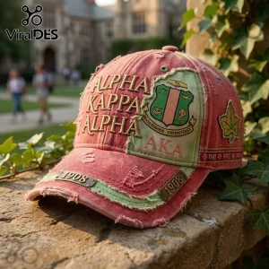 Alpha Kappa Alpha Pink Distressed Baseball Cap with gold embroidery, AKA crest, and 1908 metal accents on a stone ledge.