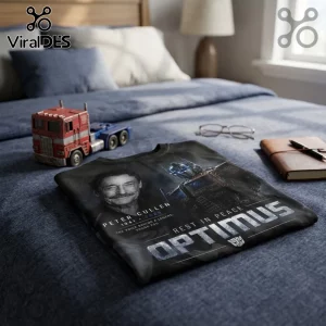 Peter Cullen Optimus Prime 1941-2026 Tribute T-Shirt featuring a portrait and helmet graphic on a folded black fabric.