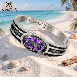 Ozzy Osbourne Prince of Darkness gold tone toggle bracelet featuring a purple gem, bat logo, and cross accents on sand.