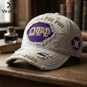 Omega Psi Phi 1911-2026 Distressed Baseball Cap in beige with purple embroidery and Greek letters on a wooden background.