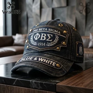 Phi Beta Sigma 1914 Denim Baseball Cap featuring dark blue fabric, gold rivets, and white fraternity embroidery on a desk.