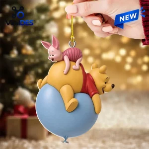 Hand holding a Winnie the Pooh and Piglet ornament featuring the pair on a blue balloon against a blurred holiday background.