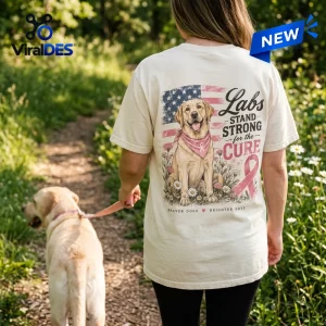 Back view of a person wearing a cream shirt with a Labrador breast cancer awareness design walking a yellow lab outdoors.