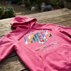 Kansas City Chiefs Crucial Catch pink hoodie with multi-colored team graphics and cancer awareness text on wooden table.