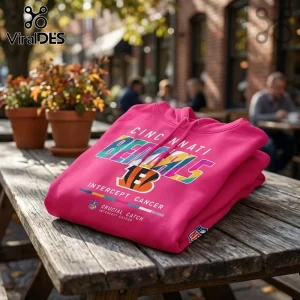 Cincinnati Bengals Crucial Catch Intercept Cancer hoodie in bright pink with multi-colored team graphics on a wooden table.