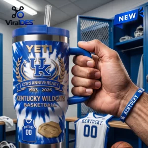 Hand holding a Kentucky Wildcats 123rd Anniversary tumbler with blue tie-dye pattern and a straw in a locker room setting.