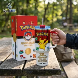Hand holding a Pokémon 30th Anniversary 40oz Stanley Tumbler next to its gift box on a wooden picnic table outdoors.