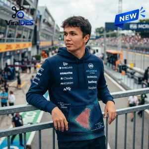 Man wearing a blue Williams F1 Team Hoodie while leaning on a metal railing near a racetrack.
