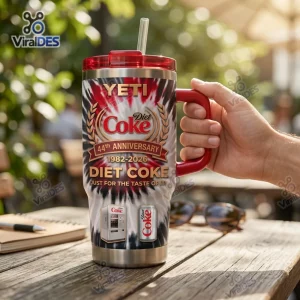 Hand holding YETI Diet Coke 44th Anniversary Handle Tumbler on outdoor wooden table.
