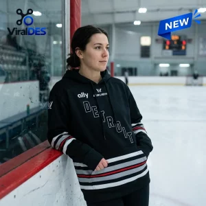 Woman wearing a PWHL Detroit Hockey 2026 black hoodie leaning against an indoor ice rink glass barrier.