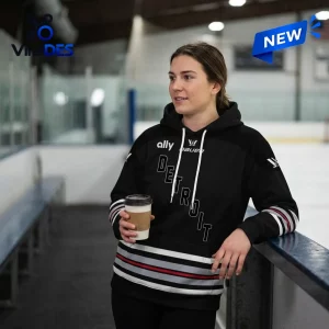Woman wearing a PWHL Detroit Hockey 2026 black hoodie holding a paper coffee cup inside an arena.