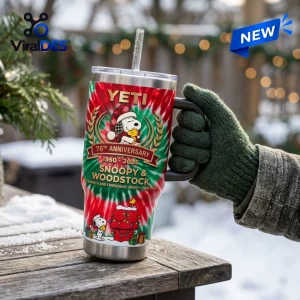 Hand in green knitted glove holding a festive YETI Stanley Tumbler with Snoopy 76th Anniversary design on a snowy table.
