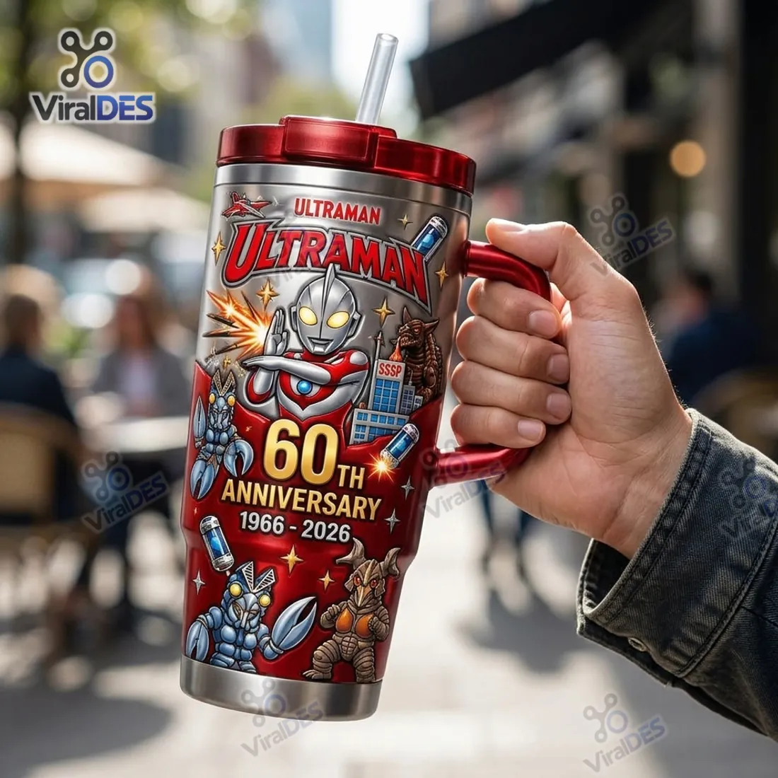Hand holding the Ultraman 60th Anniversary 1966-2026 Tumbler Cup by its crimson side handle displaying Alien Baltan and Zetton artwork
